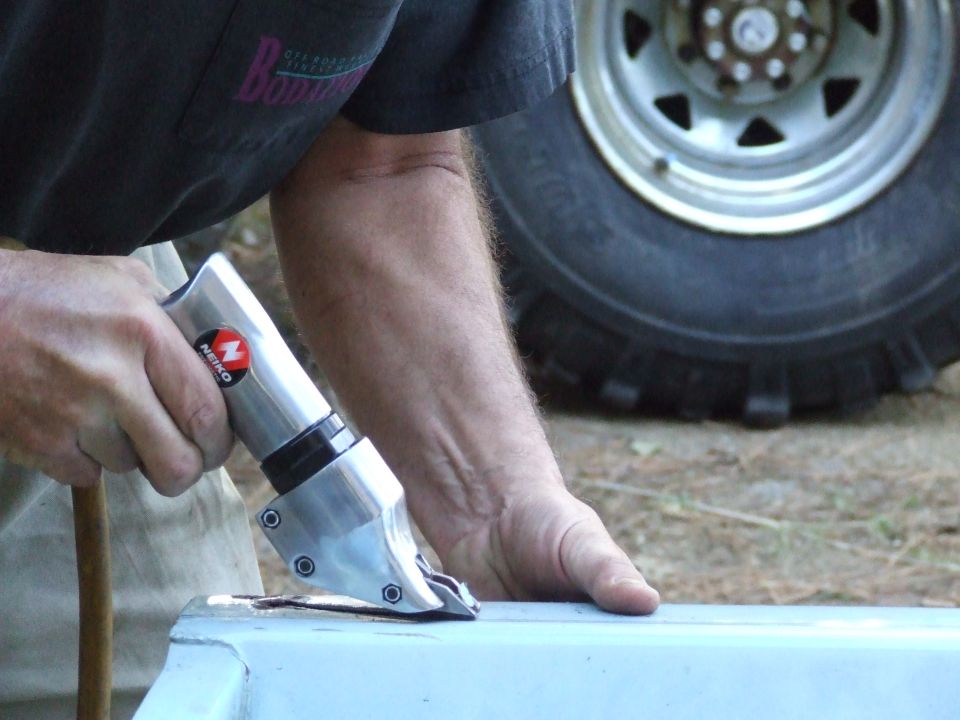 Close-up of hands using a metal shear tool on a light blue panel, with a large tire in the background.