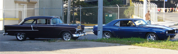 Two classic cars, a dark purple sedan and a blue coupe, parked side-by-side.