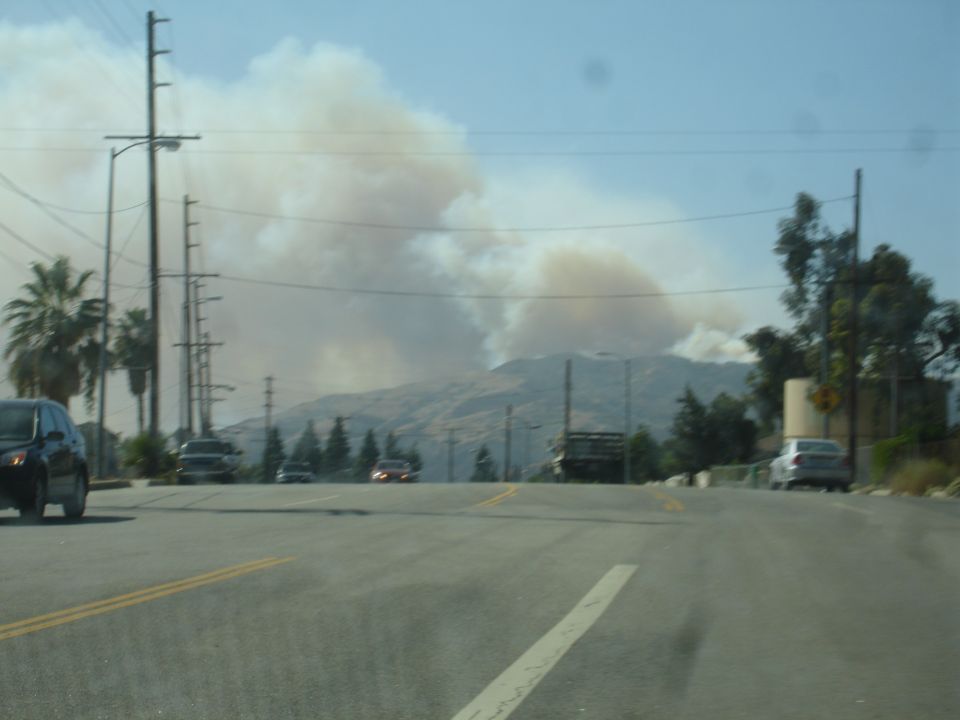Black sedan parked on the side of a road with smoke rising from hills in the background.