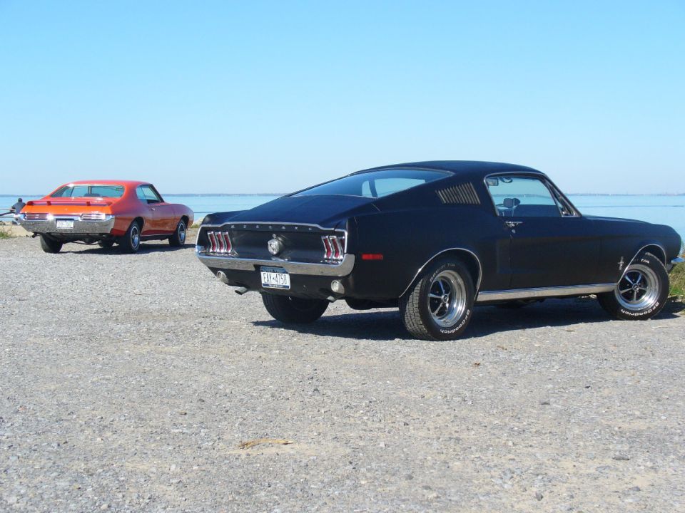 Black 1968 Ford Mustang fastback parked on gravel lot with orange classic car and water in background.