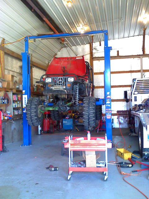 1992 Ford Bronco - A modified off-road vehicle is suspended on a lift inside a workshop, with tools and another vehicle visible nearby.