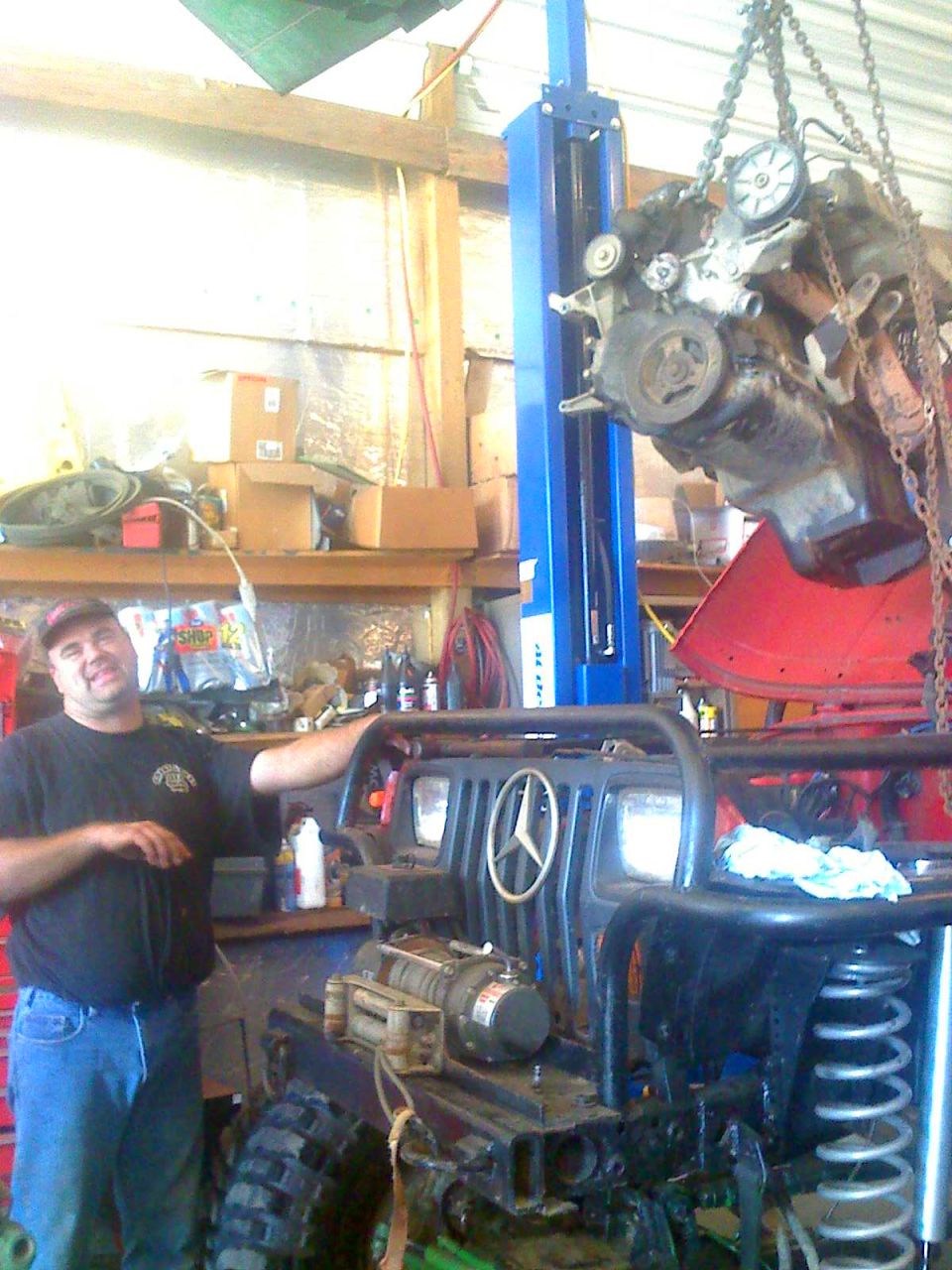 Photo of a man in a shop beside a Jeep with an engine on a hoist