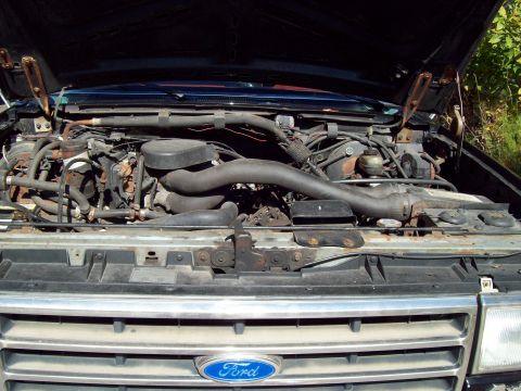 1989 Ford F-150 - The open hood of a 1989 Ford F-150 reveals a dusty engine bay with various hoses, wires, and engine components visible. The blue Ford oval…