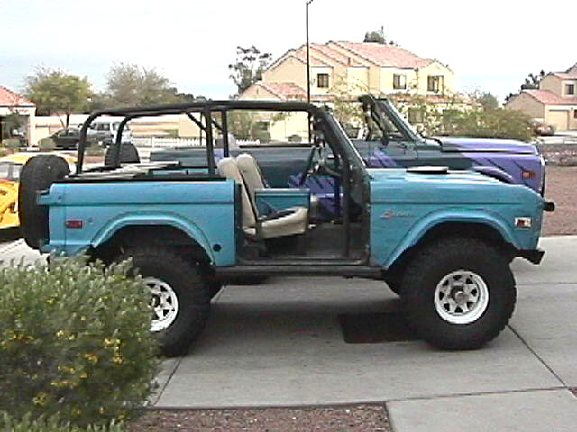 Side view of teal early Ford Bronco, doors and top off, roll cage, white wheels with large off-road tires