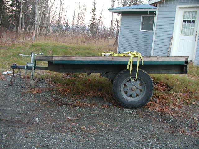 Side view of a single-axle wooden flatbed utility trailer parked on gravel by a house.