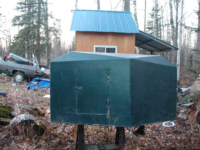 Home-built green plywood trailer cargo box with rear door, on stands in a wooded yard