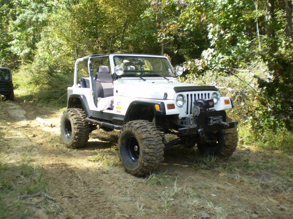 White Jeep Wrangler with off-road tires and winch on a dirt path.