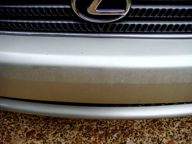 Close-up of silver Lexus front bumper below chrome badge, showing dirt band on paint