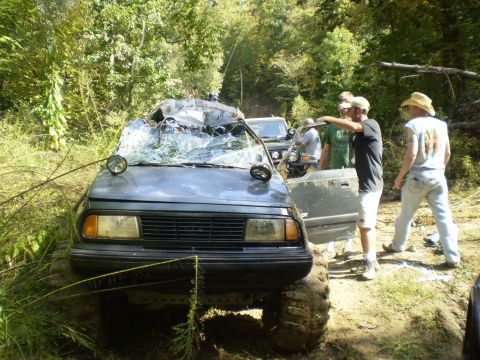 1997 Jeep Wrangler - A heavily damaged dark colored SUV with a shattered windshield is stuck in the mud with several people standing around it. The driver's sid…