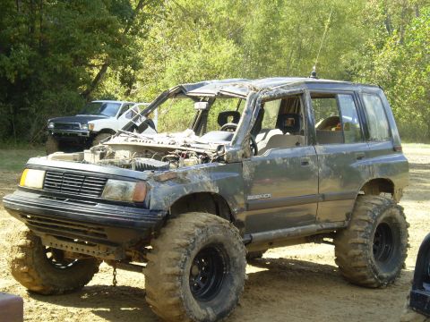 1997 Jeep Wrangler - A heavily modified, dark gray Suzuki Sidekick is parked on a dirt path with its hood open. The vehicle has oversized tires and appears to h…