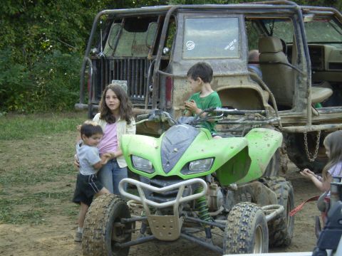 1997 Jeep Wrangler - A young girl stands next to a green ATV while a boy sits on it. A modified Jeep Wrangler is parked behind them in a muddy outdoor setting.