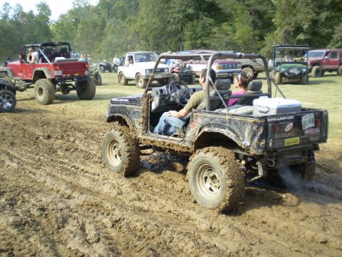 1997 Jeep Wrangler - A black Suzuki Samurai is driving through thick mud with two people inside. Other off-road vehicles are visible in the background on a sunn…