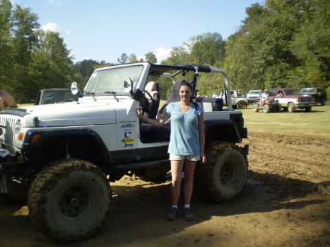 1997 Jeep Wrangler - A woman stands next to a white Jeep Wrangler with large, muddy tires. Several other vehicles, including a maroon pickup truck and an ATV, a…
