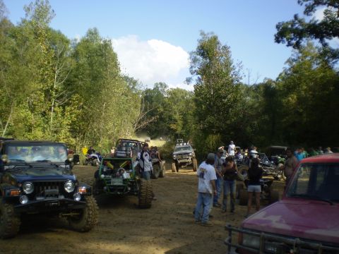 1997 Jeep Wrangler - A group of people stand around several off-road vehicles, including a black Jeep Wrangler, on a dirt path surrounded by trees.