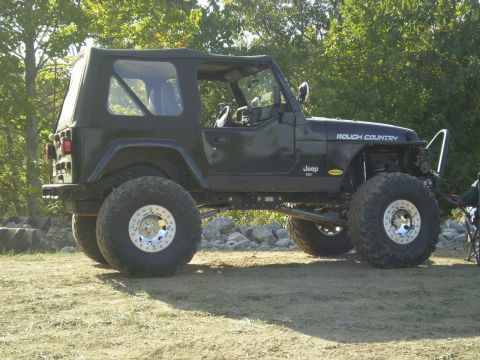 1997 Jeep Wrangler - A black Jeep Wrangler with a soft top is parked on a dirt and rocky surface, with trees in the background. The vehicle features large off-r…