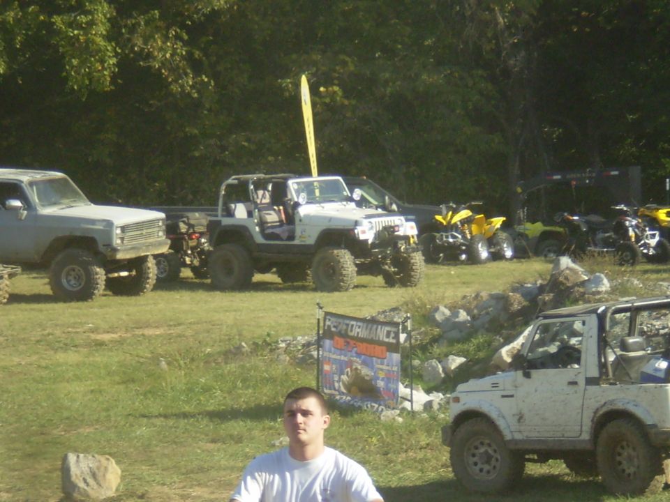 White Jeep Wrangler parked on grass with other off-road vehicles and ATVs in the background.