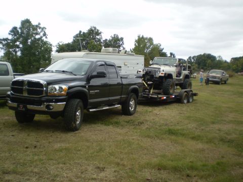 1997 Jeep Wrangler - A black Dodge Ram 2500 pickup truck tows a trailer carrying a white Jeep Wrangler. The truck is parked on a grassy field with a white trail…