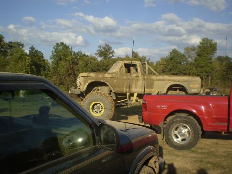 1997 Jeep Wrangler - A lifted, mud-covered vintage pickup truck with large yellow-rimmed tires is parked next to a bright red pickup truck. The scene is outdoor…