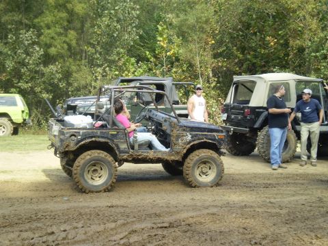 1997 Jeep Wrangler - A group of off-road vehicles are gathered in a muddy terrain, with people standing around and one person driving a black vehicle.