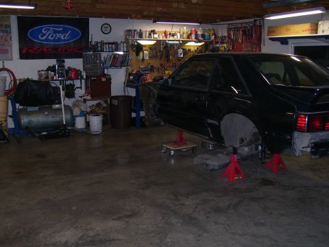 1990 Ford Mustang - A black coupe, possibly a 1990 Ford Mustang, is in a garage with its wheels removed and body panels missing.