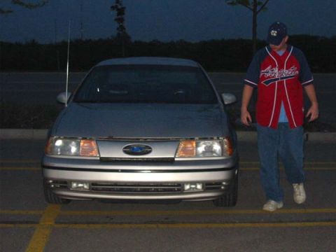 1990 Ford Taurus - A silver sedan is parked in a lot at dusk, with a person in a baseball jersey walking past it.