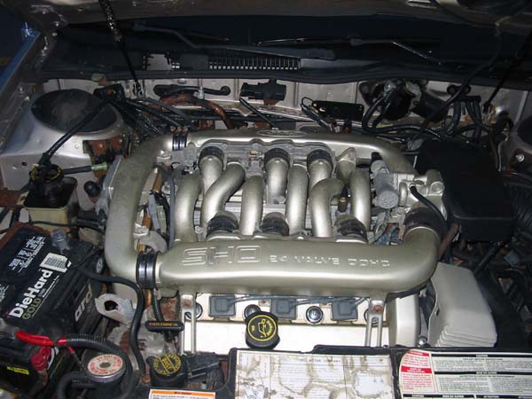 Engine bay of a 1990 Ford Taurus SHO, showing the 3.0L V6 engine with its distinctive intake manifold and a DieHard Gold battery
