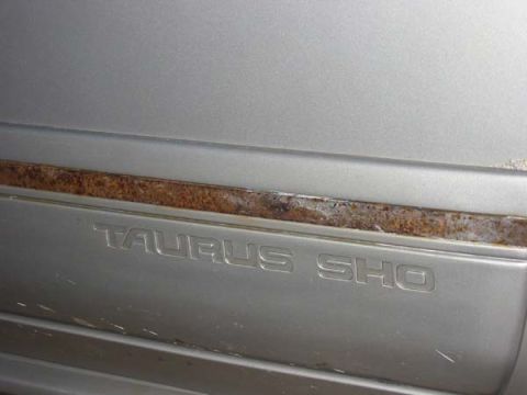 1990 Ford Taurus - Close-up of the Taurus SHO badge on a silver car, with visible rust on the trim above.