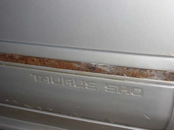 Close-up of "TAURUS SHO" badge on silver car body, showing rust on trim above