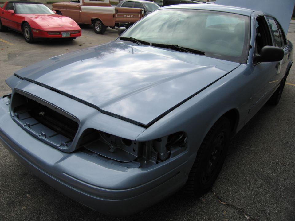 Light blue 2001 Ford Crown Victoria with hood open, missing grille and headlights. Red Corvette and orange pickup truck in background