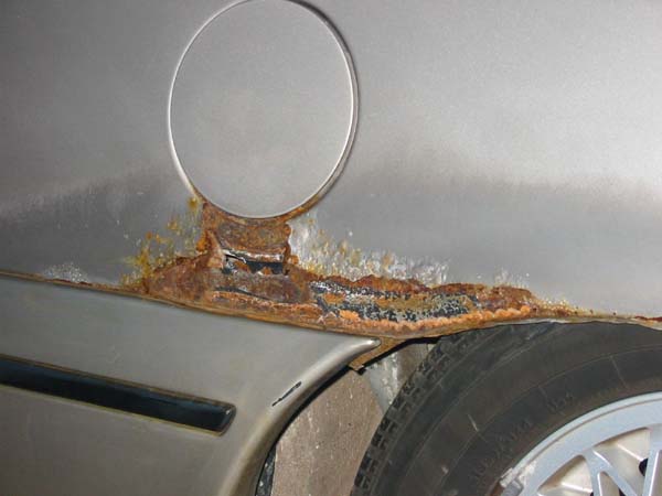 Silver car body panel with significant rust around the fuel door and lower edge, showing a tire and part of a black trim piece