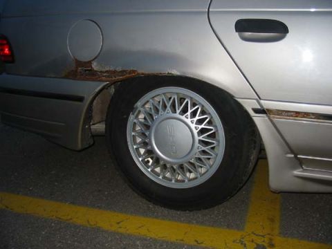 1990 Ford Taurus - Detail of a silver car's rear quarter panel, showing significant rust around the wheel well and a distinctive multi-spoke wheel.