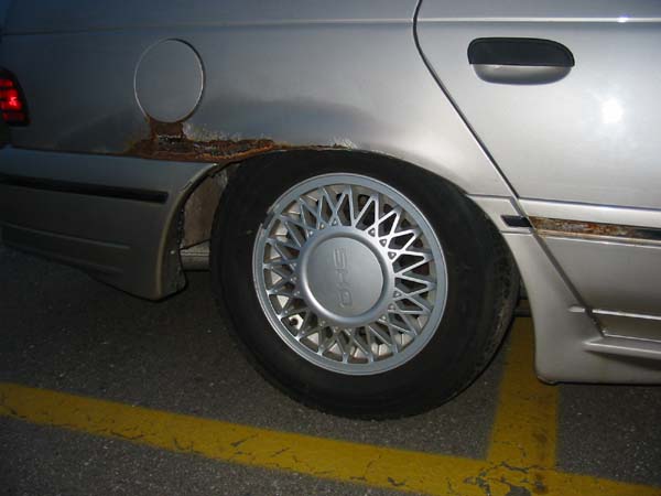 Silver car's rear wheel well with rust and a multi-spoke wheel