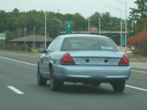 2001 Ford Crown Victoria - Rear view of a light blue sedan on a highway, with a building and trees in the background.