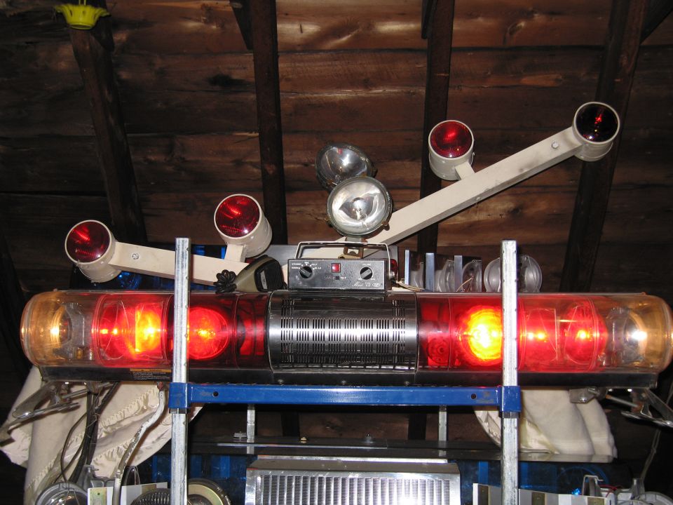 Emergency light bar with red and yellow lights, two spotlights, and control box, mounted on a blue rack in an attic