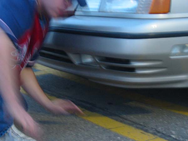 Person reaching towards front bumper of a silver car. Yellow lines on asphalt