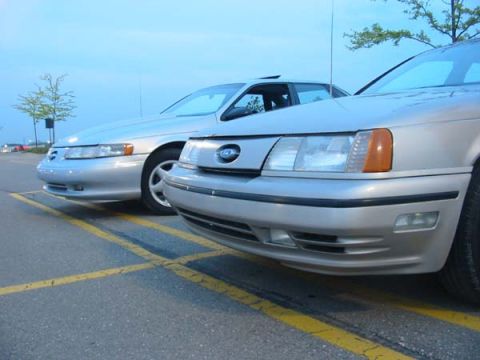 1990 Ford Taurus - Two silver sedans are parked side-by-side in a parking lot under a cloudy sky.