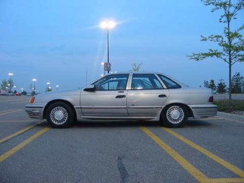 1990 Ford Taurus - This 1990 Ford Taurus sedan is captured in a side profile view while parked in a lot under a bright sky.