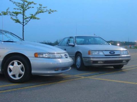 1990 Ford Taurus - Two silver sedans are parked in a lot under a clear blue sky, with a tree visible in the background.