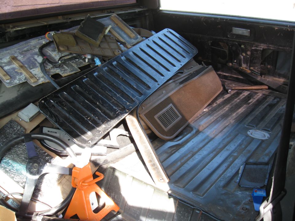 Interior car parts piled in a truck bed, including a black grille and a tan panel with a vent. An orange jack stand is visible