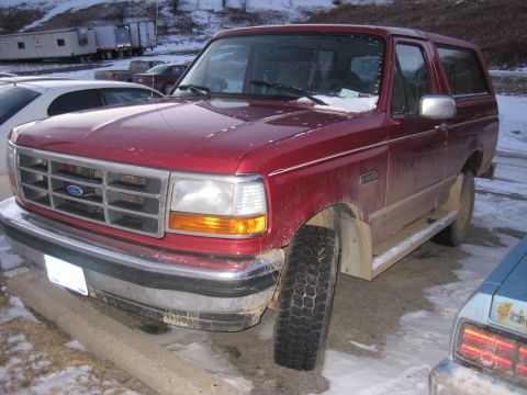 1994 Ford Bronco - A red Ford Bronco is parked in a lot with snow on the ground, showing its front grille and side.