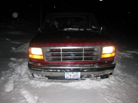 1994 Ford Bronco - A red 1994 Ford Bronco is stuck in deep snow at night, with its headlights on.