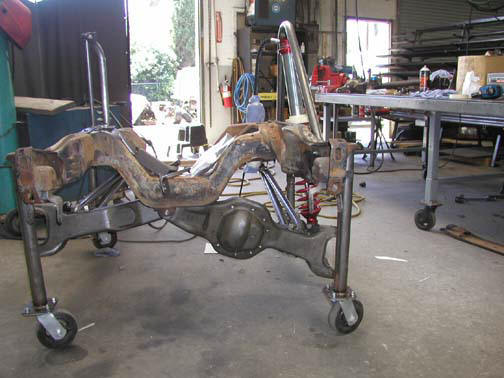 Rusty vehicle chassis with suspension components and a rear axle, mounted on a rolling stand in a workshop