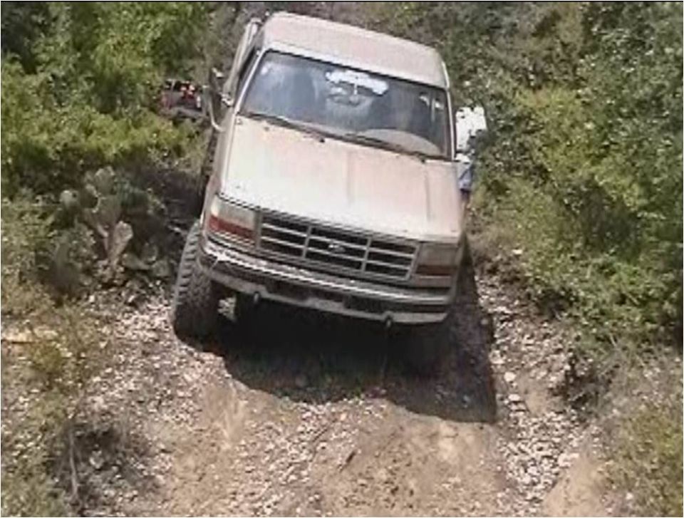 Tan Ford Bronco driving off-road on a dirt path with trees and brush on either side. Another vehicle is visible in the background