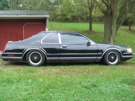 1996 Ford Bronco - The side profile of a black two-door coupe with chrome trim and aftermarket wheels is parked on a grassy lawn in front of a red shed.