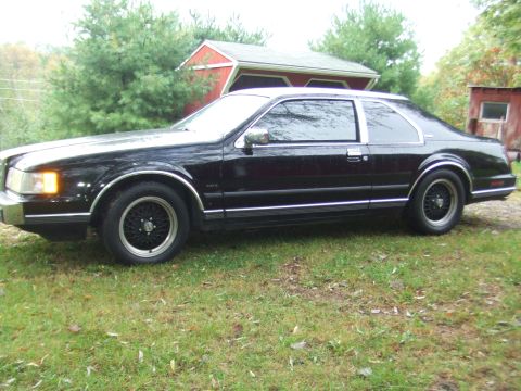 1996 Ford Bronco - A black coupe with chrome trim sits parked on a grassy lawn in front of a red barn and trees. The car has wire-spoke wheels and a silver ro…