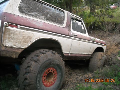 1979 Ford Bronco - A mud-covered, maroon and white 1979 Ford Bronco is stuck in thick mud on an off-road trail. The vehicle has large, aggressive tires and ap…