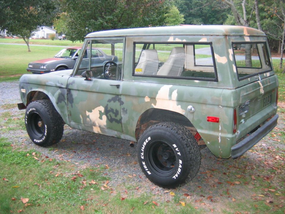 Camouflage-painted vintage Ford Bronco SUV, rear 3/4 view on gravel