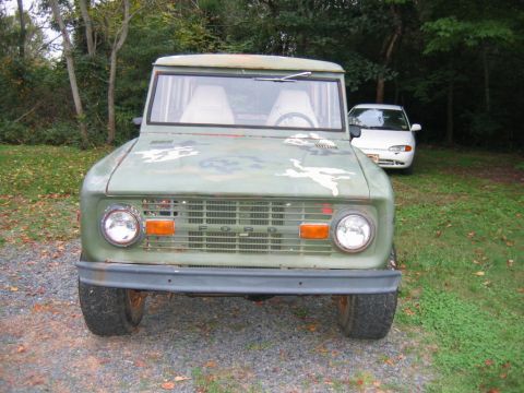 1992 Ford Mustang - Head-on view of a weathered green early Ford Bronco parked on grass and gravel at a wooded lot, its hood and fenders showing patchy primer…