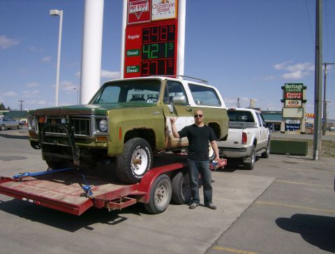 2000 Ford F-250 - A man stands next to a 1973 Chevrolet K5 Blazer being towed on a trailer, with a gas station and other vehicles in the background.