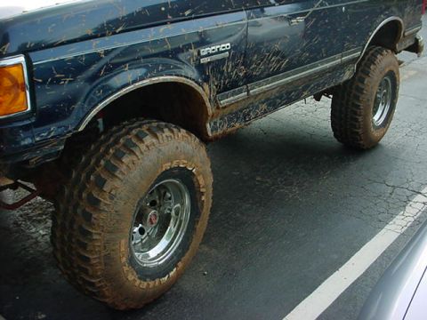 1989 Ford Bronco - A muddy dark blue Ford Bronco is shown from the side, highlighting its large tires and off-road condition.